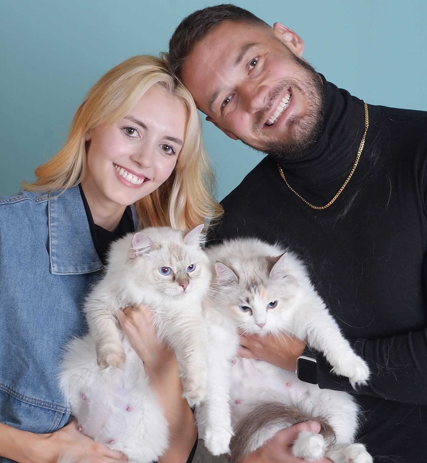 Two smiling people against a teal backdrop, each holding a long-haired cream Ragdoll cat with blue eyes